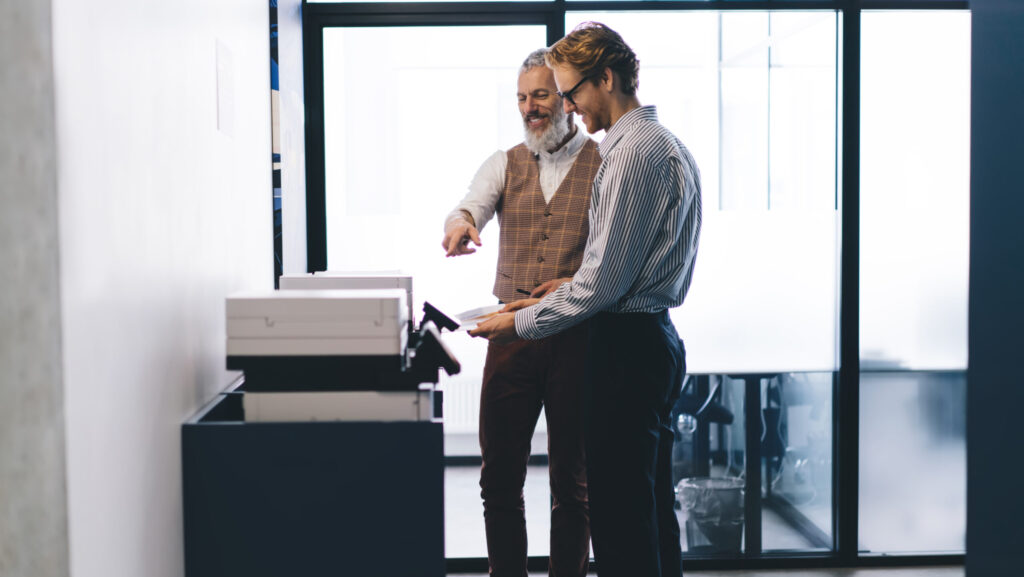 Two men standing by an office multifunction printer, smiling 