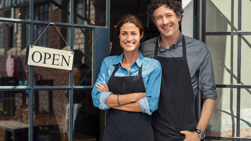 picture of a woman and man smiling in front of a store with an open sign.