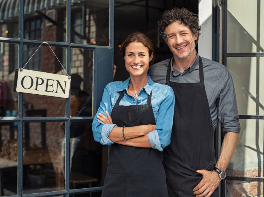 picture of a woman and man smiling in front of a store with an open sign.